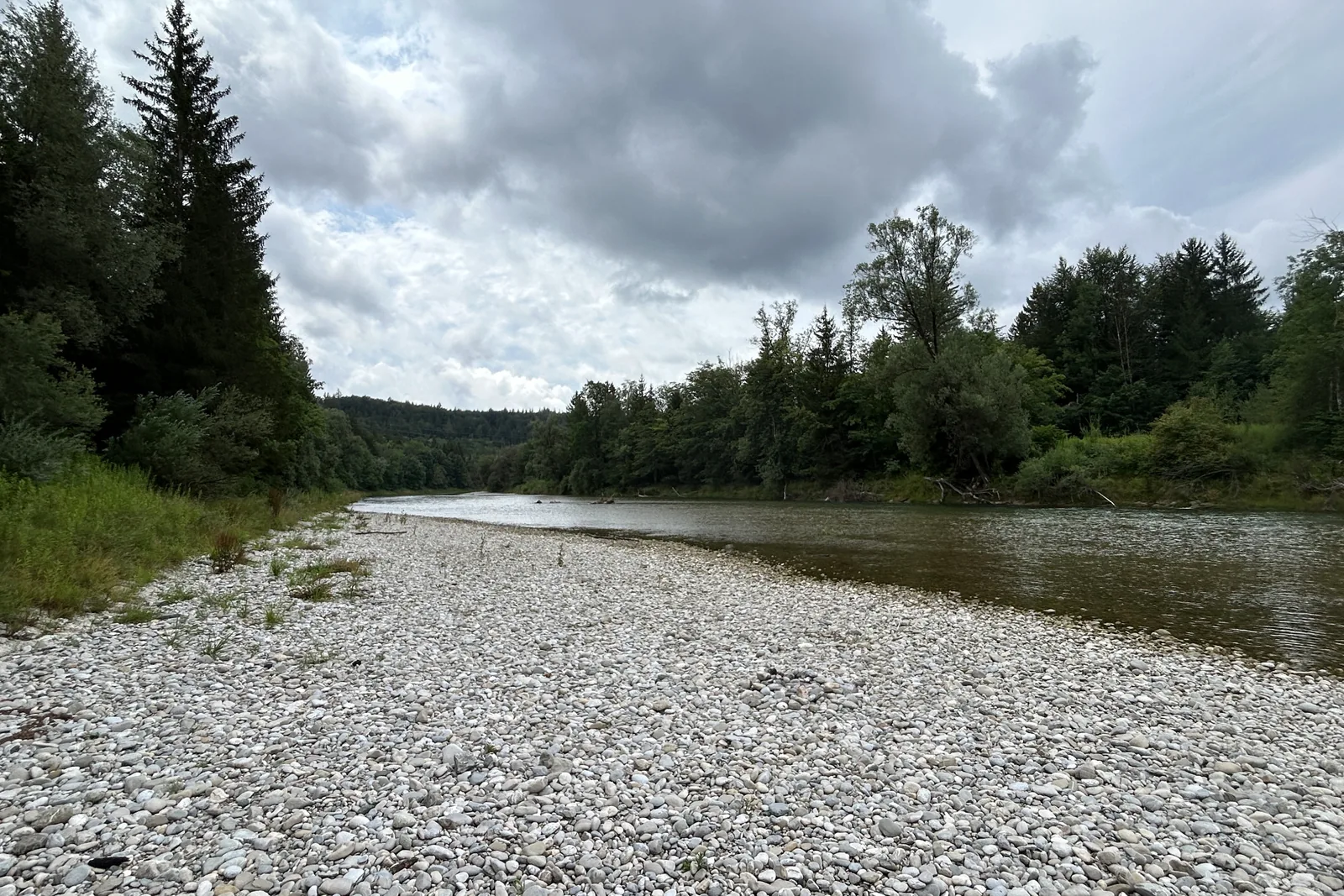 Breites Kiesufer an der Isar bei Grünwald mit bewaldeten Hängen und dunklen Wolken